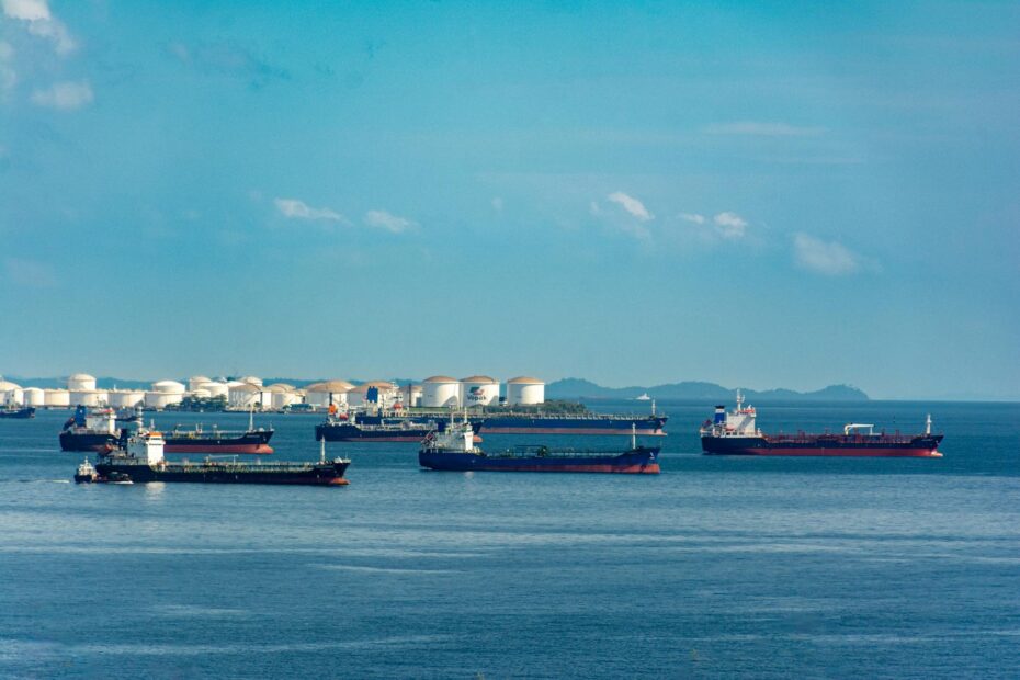 Oil markets and geopolitical risk — A fleet of cargo ships docked near oil storage tanks along a serene coastline with a clear blue sky above.